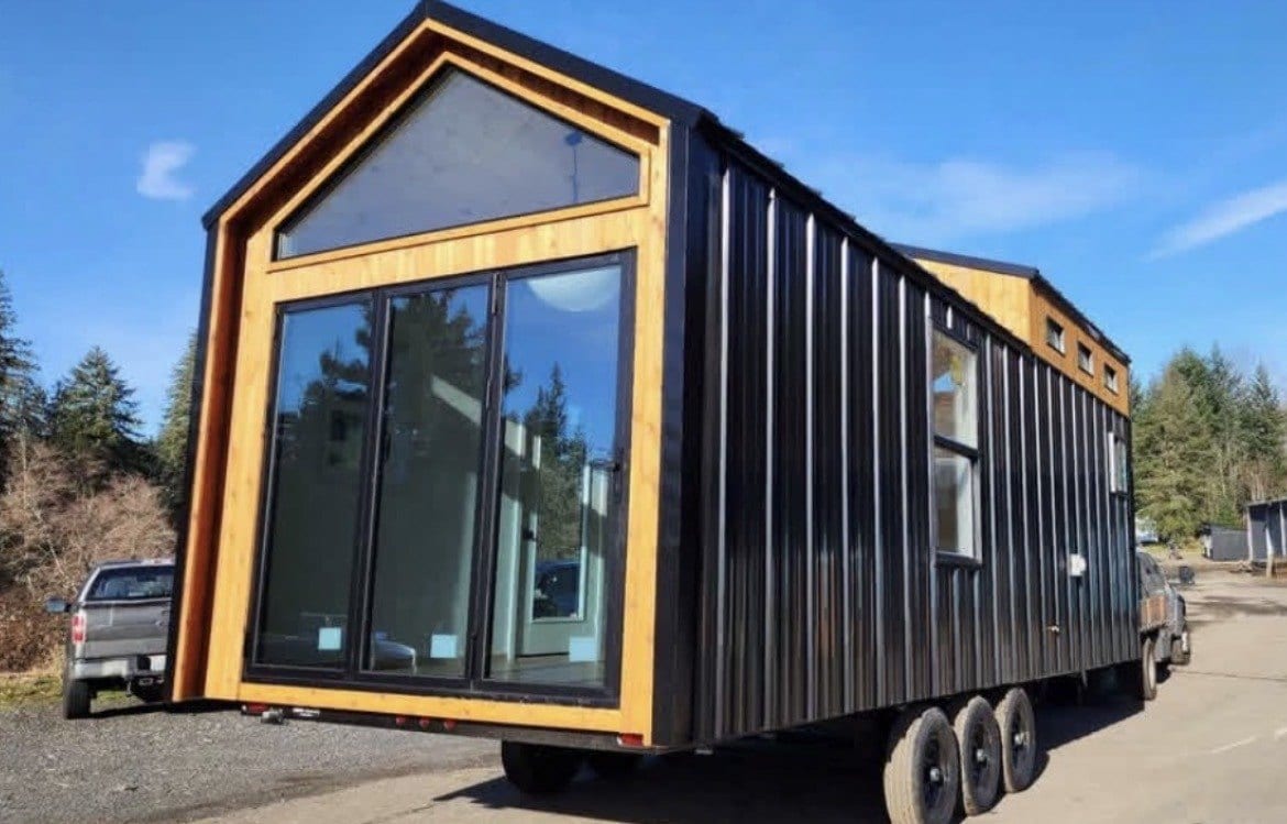 Tiny home in black metal siding with a cedar-framed glass gable end, on a triple-axle trailer